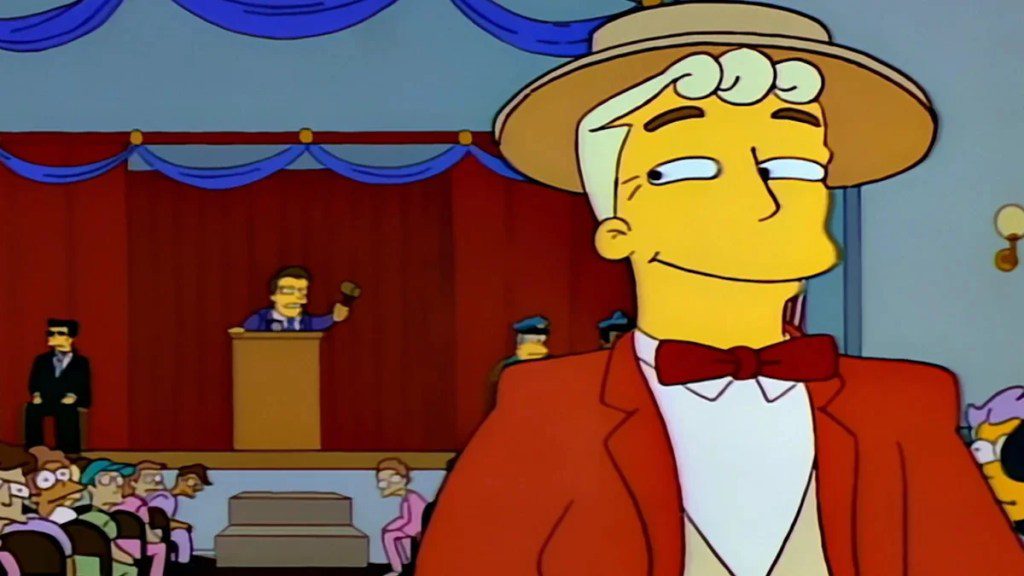 A smiling man in a red jacket, bowtie and straw hat looks smugly over his shoulder at a town hall meeting in a scene from animated series "The Simpsons."