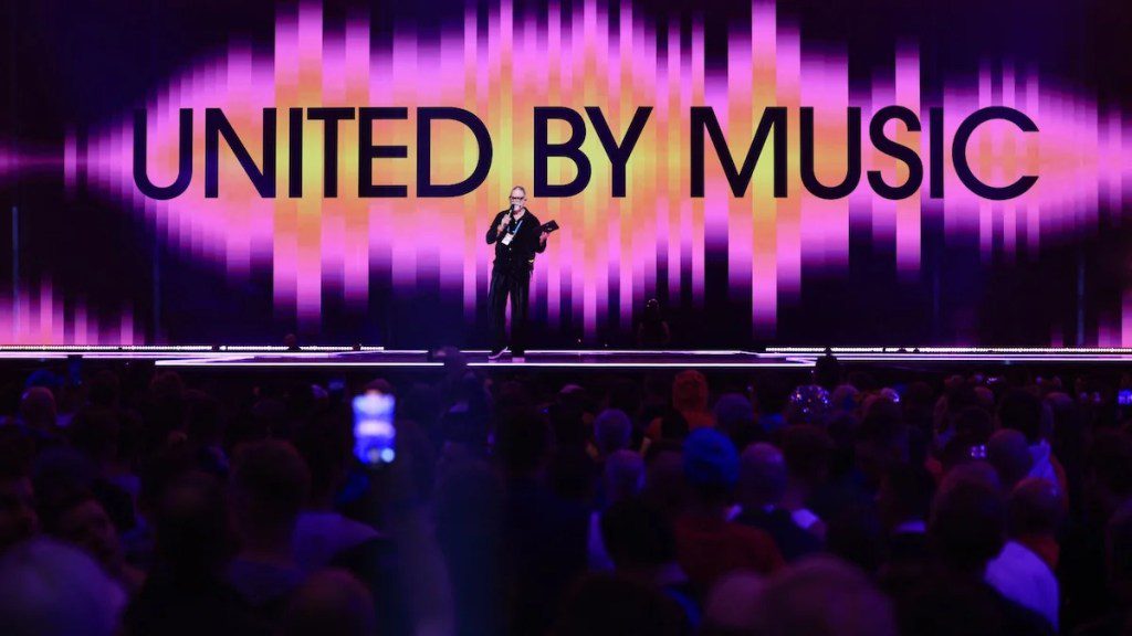 A man on stage in front of many people, with a graphic behind reading "UNITED BY MUSIC."